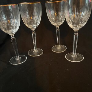 Puede incluir: Cuatro copas de vino transparentes con bordes dorados. Las copas tienen un diseño grabado detallado en el cuenco y un tallo delgado. Las copas están dispuestas sobre una superficie oscura.