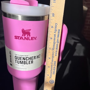 May include: A pink Stanley Quencher H2.0 tumbler with a white lid and straw. The tumbler has a handle and the Stanley logo. The label reads "THE FLOWSTATE QUENCHER H2.0 TUMBLER 40 OZ | 1.18L All Day Hydration". The tumbler is approximately 20 cm tall.