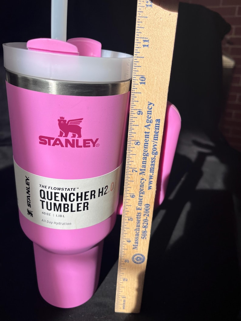 May include: A pink Stanley Quencher H2.0 tumbler with a white lid and straw. The tumbler has a handle and the Stanley logo. The label reads "THE FLOWSTATE QUENCHER H2.0 TUMBLER 40 OZ | 1.18L All Day Hydration". The tumbler is approximately 20 cm tall.