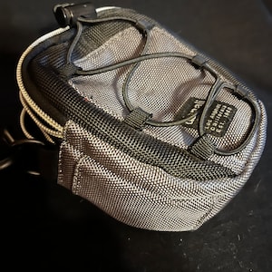 Puede incluir: Una pequeña bolsa de cordón gris y negro con una correa negra y una cremallera negra. La bolsa tiene una etiqueta que dice "Carhartt, Los Ángeles, California, Est. 1989".