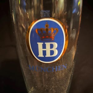 May include: A clear beer glass with a blue and gold logo featuring a crown and the letters "HB". The word "MUNCHEN" is printed in blue below the logo. The glass is tapered and has a long stem.