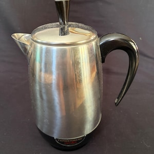May include: A stainless steel Farberware percolator coffee pot with a polished metallic finish, a black plastic handle, and a matching black knob on the lid. The base features a black circular accent with the Farberware brand name printed in white.