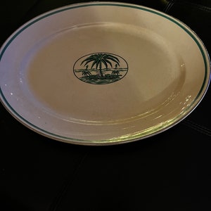 Puede incluir: Una fuente ovalada con una superficie blanca y un borde verde. El centro presenta un diseño de palmera verde dentro de un marco ovalado. La fuente probablemente está hecha de cerámica o porcelana.