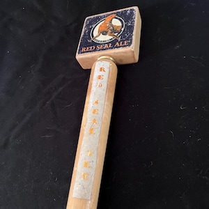 Puede incluir: Un tirador de cerveza de madera con una parte superior cuadrada que muestra un gráfico de una foca y el texto RED SEAL ALE. El mango cilíndrico tiene una etiqueta blanca vertical con el texto naranja RED SEAL ALE sobre madera clara.