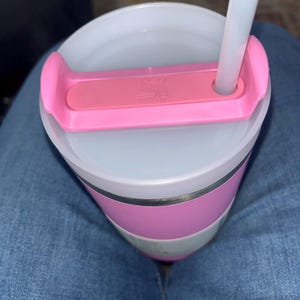 May include: A pink and white insulated tumbler with a clear lid and a pink handle. A white straw is inserted into the lid. The tumbler has a pink band around the middle and a silver band below it.