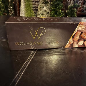 May include: A black box with gold lettering that says "Electric Wine Opener" and "Wolfgang Puck". The box has a gold "W" logo and a close-up of wine corks on the right side.