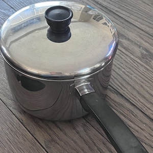 May include: A stainless steel saucepan with a lid and a black handle. The pot has a shiny, reflective surface and a black knob on the lid. The handle is attached to the side of the pot. The pot is on a wooden surface.