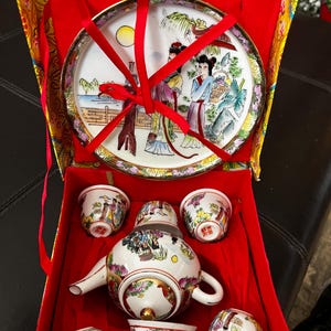 May include: A white porcelain tea set with a red and gold box. The tea set includes a teapot, six cups, and a plate with a painted scene of women in traditional clothing.