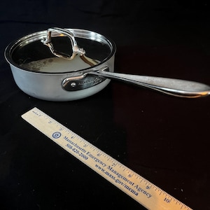 Pode incluir: Panela de aço inoxidável com tampa de vidro e cabo longo. A panela tem um acabamento prateado e uma tampa de vidro transparente. Uma régua de madeira com texto está em primeiro plano. A panela tem aproximadamente 15 cm de diâmetro.