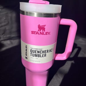 May include: A pink Stanley Quencher H2.0 tumbler with a handle and a white lid. The tumbler has a straw and a label that reads "THE FLOWSTATE QUENCHER H2.0 TUMBLER 1.18 L All Day Hydration". The Stanley logo is visible.