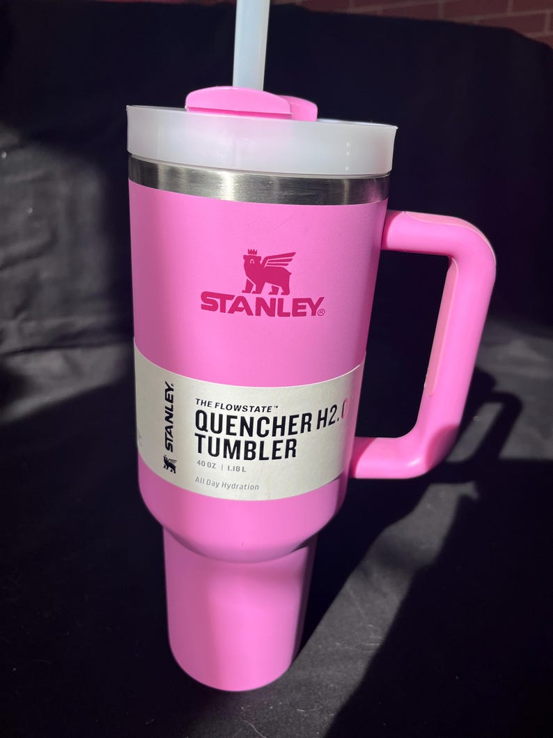 May include: A pink Stanley Quencher H2.0 tumbler with a handle and a white lid. The tumbler has a straw and a label that reads "THE FLOWSTATE QUENCHER H2.0 TUMBLER 1.18 L All Day Hydration". The Stanley logo is visible.