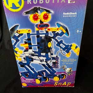 May include: A Robotix Snap sound-activated robot building set. The box features a robot design with blue and yellow components, large yellow eyes, and the text "Robotix" and "RadioShack Exclusive!". The box states "Build 3 different sound-activated motorized robots!"