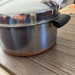 Puede incluir: Una olla de acero inoxidable con base de color cobre, que incluye una tapa de acero inoxidable a juego con un pomo negro y un asa lateral negra. La superficie de metal pulido refleja claramente el entorno circundante.