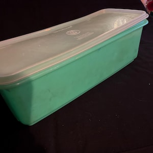 Puede incluir: Un recipiente rectangular verde claro para guardar alimentos con una tapa transparente ajustada. El recipiente está hecho de plástico duradero y está diseñado para guardar alimentos. La tapa tiene un borde blanco y un logotipo. Sobre una superficie negra.