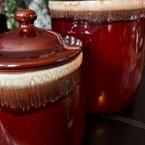 以下が含まれることがあります： ふた付きのセラミック製の容器が2つ。容器は茶色で、上部に白い帯があります。容器は暗い表面の上に置かれています。