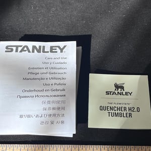 May include: A product information sheet and a small card with the Stanley logo and "QUENCHER H2.0 TUMBLER" text. The product information sheet has text in multiple languages, including English, Spanish, French, and German.