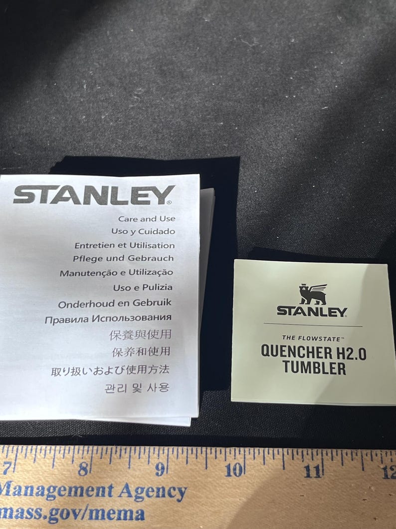 May include: A product information sheet and a small card with the Stanley logo and "QUENCHER H2.0 TUMBLER" text. The product information sheet has text in multiple languages, including English, Spanish, French, and German.