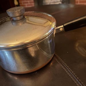 May include: A stainless steel saucepan with a copper-coloured base, a matching lid with a round knob, and a long black handle. The pot features a polished metallic finish that reflects light, and the lid fits securely over the top of the circular vessel.
