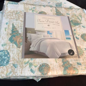 Puede incluir: Un juego de funda nórdica individual en un paquete de plástico con tela blanca y un estampado de conchas y corales en color verde azulado y bronceado. La etiqueta dice Antigua Collection Twin Duvet Set, Wrinkle Resistant, Easy Care.