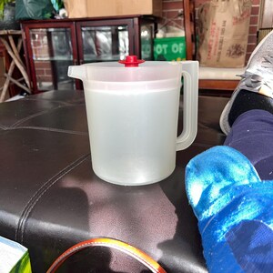 Può includere: Un'ampolla di plastica trasparente con un coperchio rosso su un pouf in pelle marrone. L'ampolla è piena di un liquido trasparente.
