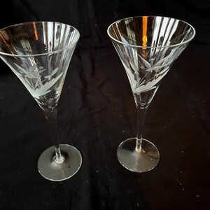 May include: A pair of clear cocktail glasses with etched floral patterns. The glasses have a conical bowl and a thin stem, set on a circular base. The etched design shows delicate leaves and stems. The background is black.