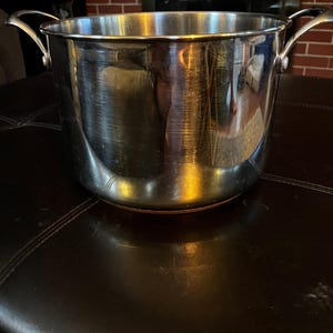 Puede incluir: Una olla de acero inoxidable con dos asas curvas. La olla tiene una superficie brillante y reflectante y se asienta sobre una superficie marrón oscuro. El fondo incluye una chimenea de ladrillo.