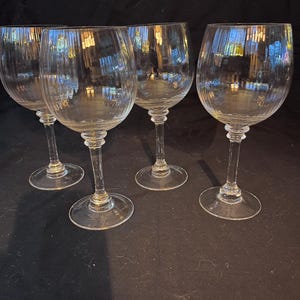 May include: Four clear, stemmed wine glasses. Each glass has a rounded bowl and a long, slender stem. The glasses are made of clear glass and are arranged on a dark surface. The glasses are designed for serving wine.