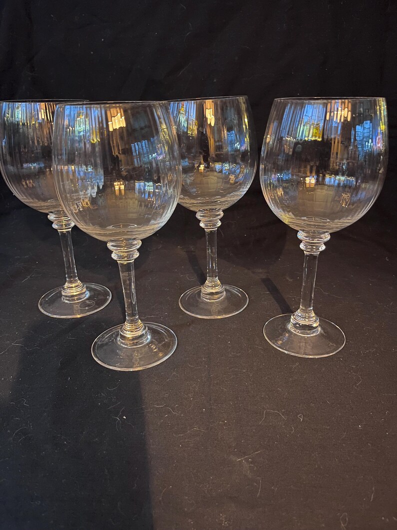 May include: Four clear, stemmed wine glasses. Each glass has a rounded bowl and a long, slender stem. The glasses are made of clear glass and are arranged on a dark surface. The glasses are designed for serving wine.