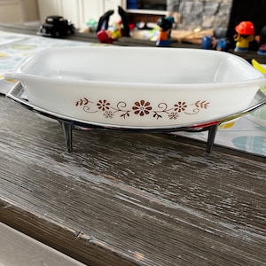 May include: White casserole dish with a brown floral design on the rim, sitting on a black metal stand.