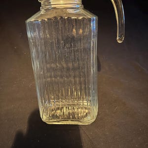May include: A clear glass jug with a handle and a white lid. The jug has vertical ribbed detailing and a rectangular shape. The handle is curved and attached to the side of the jug. The lid is flat and sits on top of the jug.
