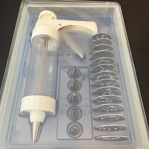 May include: A white and clear cookie press set in a clear plastic container. The set includes a cookie press, various decorating tips, and several cookie shape templates. The text "KUHN RIKON" and "Cookie Press" are visible on the press.