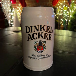 May include: White ceramic beer stein with a black and gold logo that reads "Dinkel Acker" and "Brau-Erfahrung aus 200 jähriger Familientradition".