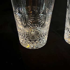 Op de afbeelding: Twee heldere, geslepen glazen drinkglazen met ingewikkelde geometrische en bloemmotieven. De glazen hebben een diamantgeslepen basis en een gedetailleerd ontwerp dat zich uitstrekt tot aan de zijkanten. Het heldere glas reflecteert licht.