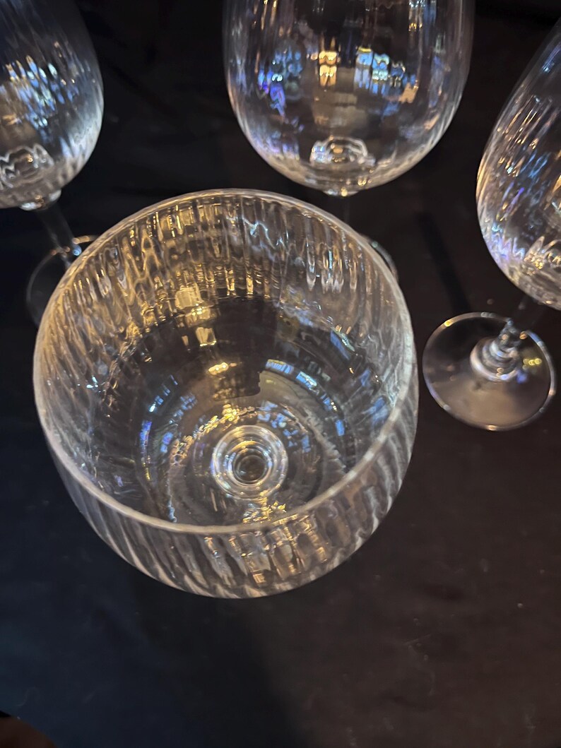 May include: A collection of clear glass stemware, including wine glasses and a large bowl. The bowl has a textured, ribbed design. The glasses are arranged against a dark background, reflecting light.