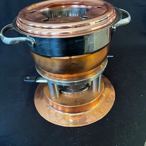 Può includere: Pentola per fondue in rame con base in acciaio inossidabile e coperchio con bruciatore incorporato. La pentola ha un'etichetta argentata con la scritta "Swissmar".