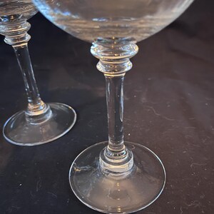 May include: Two clear stemmed wine glasses. The glasses have a round base, a long stem, and a bowl-shaped top. The stem has a decorative design near the bowl. The glasses are on a dark surface.