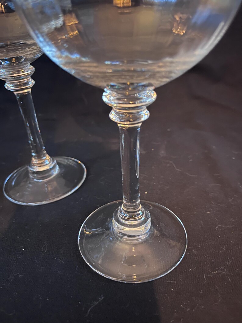 May include: Two clear stemmed wine glasses. The glasses have a round base, a long stem, and a bowl-shaped top. The stem has a decorative design near the bowl. The glasses are on a dark surface.