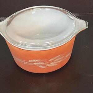 Könnte beinhalten: Eine orangefarbene Pyrex-Auflaufform mit einem Deckel aus klarem Glas. Die Schüssel hat ein Weizenmuster in Weiß und Griffe. Ein Vintage-Küchenartikel.