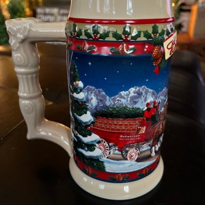 Può includere: Una boccale di birra natalizia Budweiser in ceramica con una scena invernale che presenta un carro rosso che trasporta un albero di Natale, due cavalli e un uomo con un cappotto rosso. Il boccale ha un manico bianco e un design blu, rosso e verde.