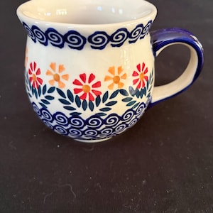 Pode incluir: Uma caneca de cerâmica com corpo arredondado e asa azul. A superfície de cor creme apresenta um padrão pintado à mão de flores cor de laranja e vermelhas com folhas verdes, delimitado por desenhos de ondas em espiral azul-escuras nas bordas.
