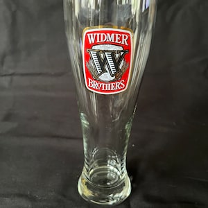 Puede incluir: Un vaso de cerveza de vidrio transparente con un logotipo rojo y dorado que dice "Widmer Brothers".
