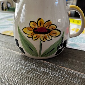 Könnte beinhalten: Weißer Keramik-Krug mit einem gelben Sonnenblumenmuster und grünen Blättern. Der Krug hat ein schwarz-weißes Schachbrettmuster am Boden und das Wort "Sweet" steht an der Seite.