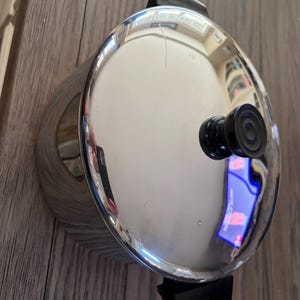 Puede incluir: Una olla de acero inoxidable con un acabado pulido y reflectante y una tapa circular. La olla cuenta con dos asas laterales de plástico negro y un pomo de plástico negro a juego en el centro de la tapa para un agarre seguro al levantar.