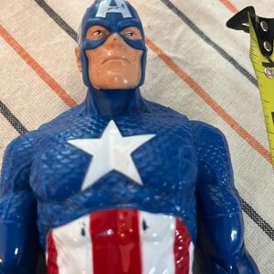 May include: A blue and red plastic action figure of Captain America. The figure has a blue and white helmet with a red star on the front. The figure is wearing a blue and red suit with a white star on the chest. The figure is holding a red, white, and blue shield.
