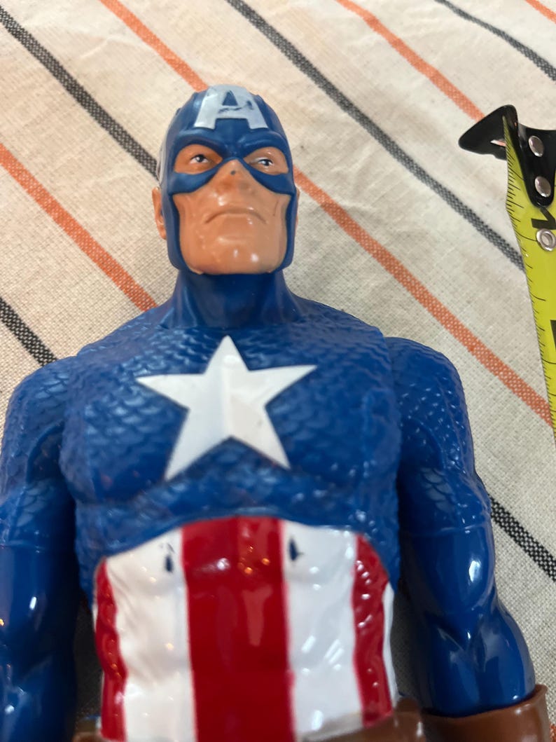 May include: A blue and red plastic action figure of Captain America. The figure has a blue and white helmet with a red star on the front. The figure is wearing a blue and red suit with a white star on the chest. The figure is holding a red, white, and blue shield.