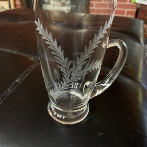 Könnte beinhalten: Eine klare Glaskaraffe mit Henkel, die mit einem Blattdesign graviert ist. Die Karaffe hat einen runden Boden und einen hohen, zylindrischen Körper.