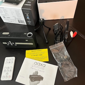 May include: A black P4H Pico Projector with a white remote, cables, and a mini tripod. The box reads "AAXA Technologies" and "P4H Pico Projector". A user guide and a yellow card with a phone number are also included.