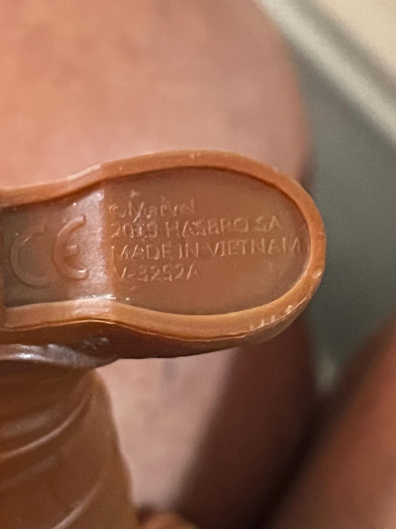 May include: Close-up of the bottom of a brown plastic toy figure. The figure has a CE mark and the text "Marvel 2019 Hasbro SA Made in Vietnam V-3252A".