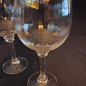 May include: Two clear wine glasses with ribbed detailing on the bowl. The glasses have a long stem and a circular base. The background is dark, and the glasses reflect the surrounding environment.