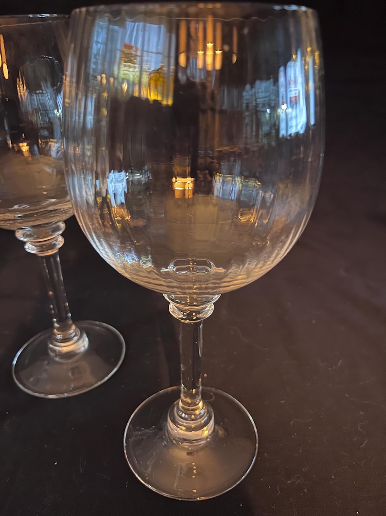 May include: Two clear wine glasses with ribbed detailing on the bowl. The glasses have a long stem and a circular base. The background is dark, and the glasses reflect the surrounding environment.